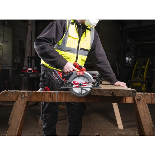 Pracownik tnie Milwaukee M18 FCS66GR3 kantówkę na kozłach, w tle maszyny stolarskie