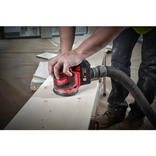 Milwaukee M18 BOS125 z podłączonym wężem odkurzacza szlifuje deskę