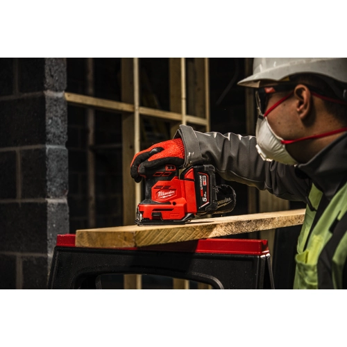 Pracownik w masce szlifuje Milwaukee M18 BQSS deskę na stole