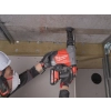 Pracownik w białym kasku wierci Milwaukee M18 FHAC16 pionowo w górę w stropie przy ruszcie sufitu