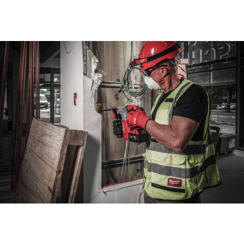 Pracownik w kasku wierci Milwaukee M18 BLH w ścianie przy oknie, obok zwinięte kable
