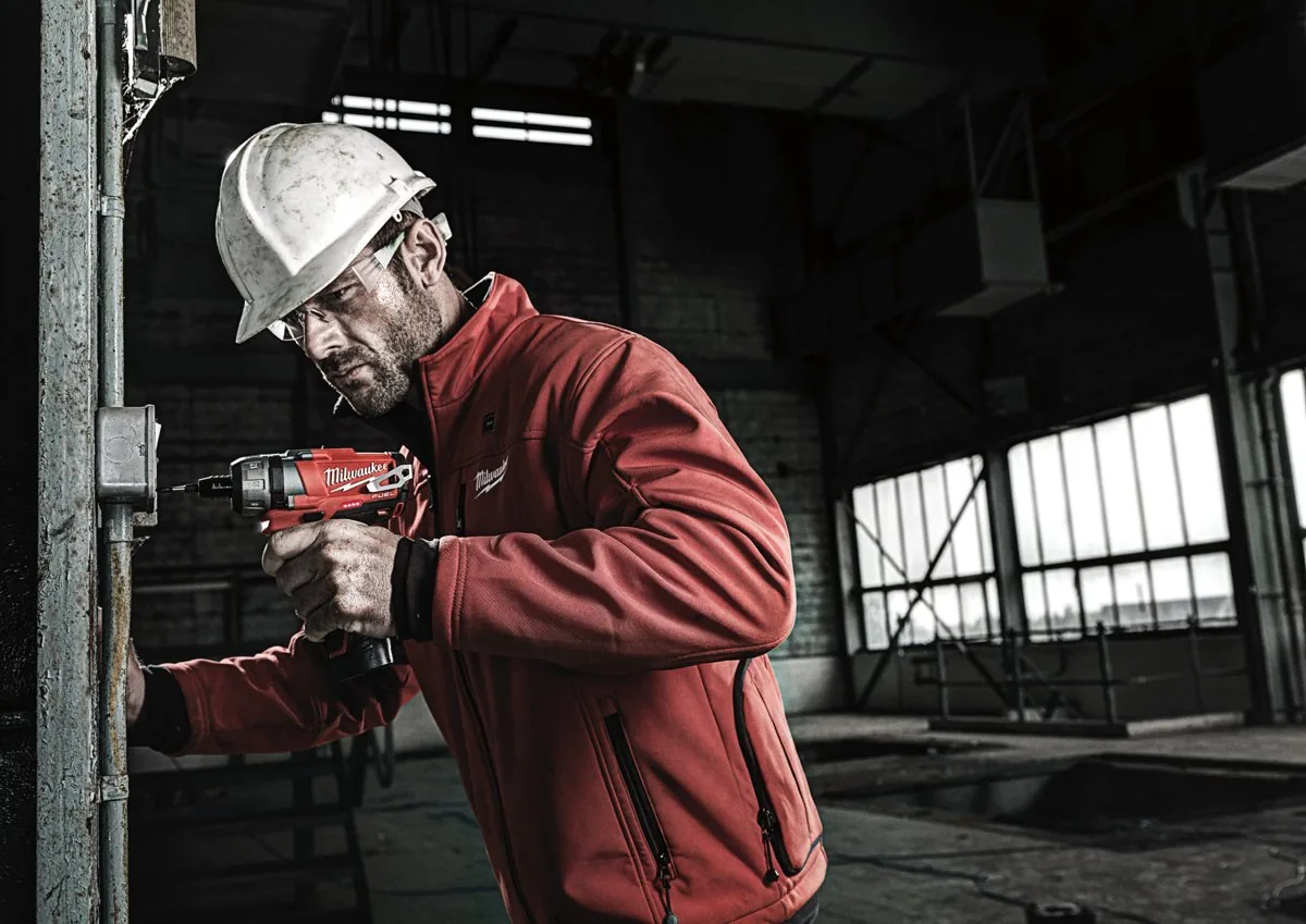 Elektryk w kasku wkręca kompaktową wkrętarką MILWAUKEE M12 przy skrzynce w hali