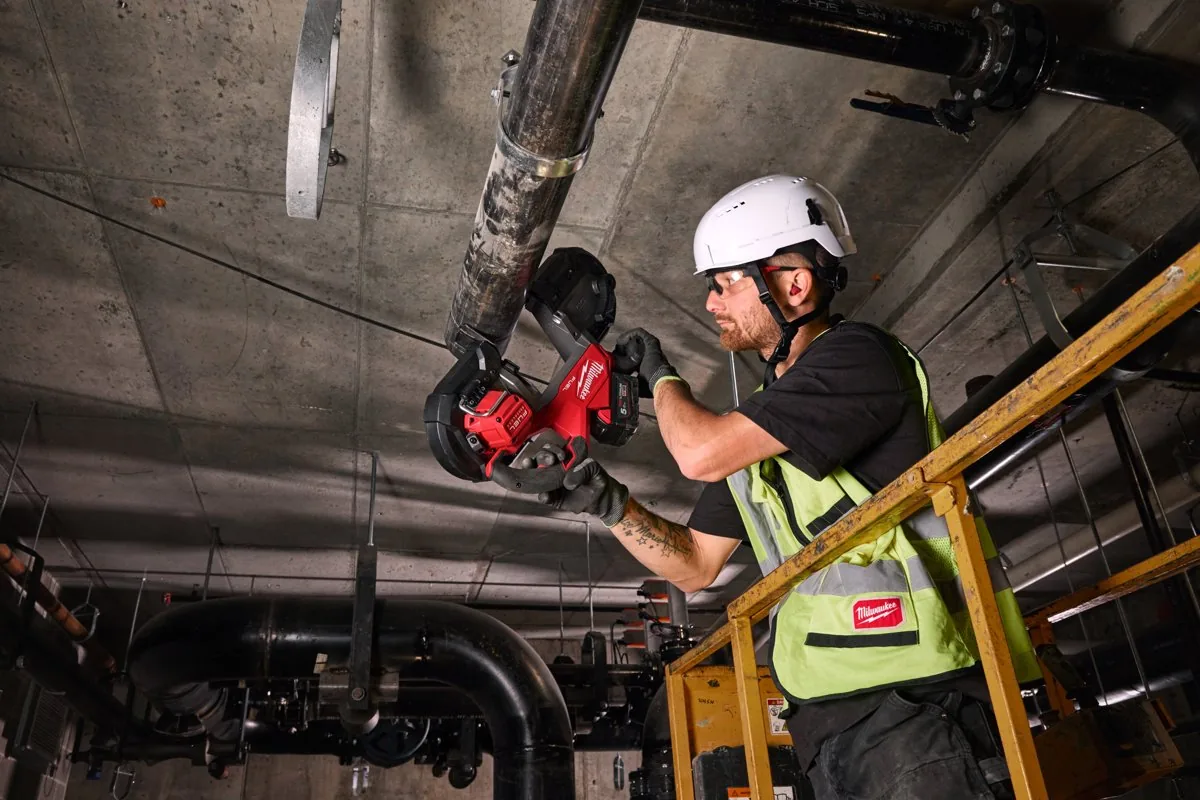 Instalator w kasku tnie M18 FBS127 rurę pod stropem piwnicy technicznej