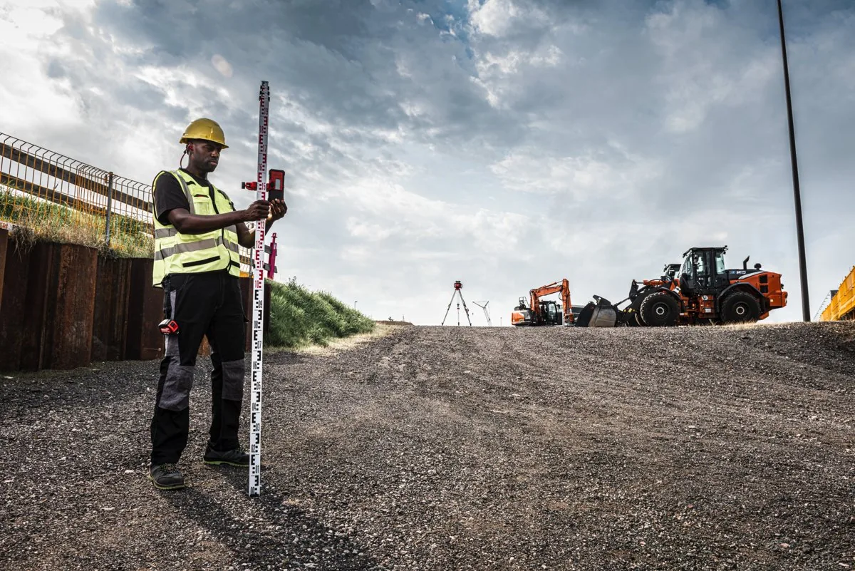 Geodeta z łatą na placu budowy przy niwelatorze M18 RLOHV1200