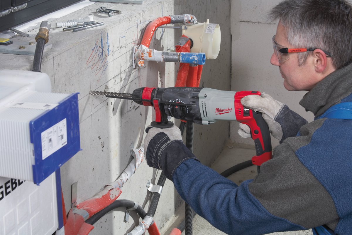 Instalator wierci sieciową młotowiertarką MILWAUKEE w betonowej ścianie przy stelażu podtynkowym